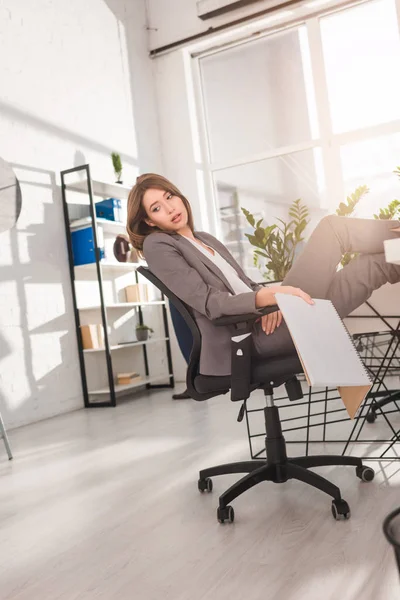 Atractiva mujer de negocios descansando en la silla y la celebración de cuaderno con la página en blanco en la oficina - foto de stock