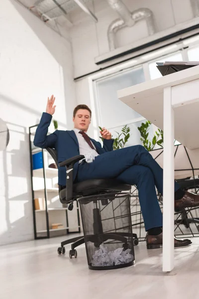 Hombre de negocios guapo sentado en la silla y tirar papel en la papelera en la oficina, concepto de dilación - foto de stock