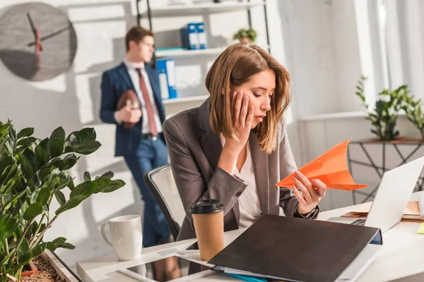 Tired businesswoman holding paper plane near laptop with coworker on background, procrastination concept — Stock Photo