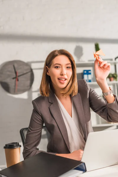 Cheerful businesswoman holding paper plane near laptop and disposable cup in office, procrastination concept — Stock Photo