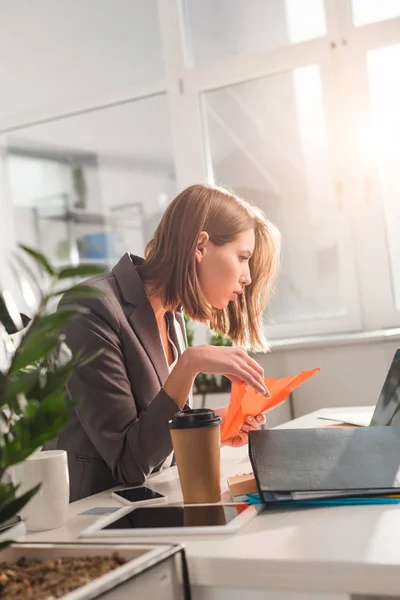 Enfoque selectivo de la mujer sosteniendo avión de papel y mirando el ordenador portátil cerca de taza desechable en la oficina, concepto de dilación - foto de stock