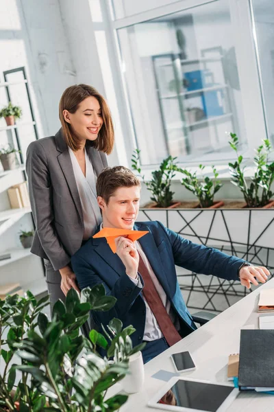 Cheerful businesswoman standing near happy coworker holding paper plane in office — Stock Photo