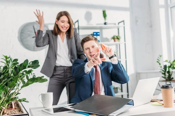 Attractive businesswoman standing and gesturing near cheerful coworker holding paper plane and pointing with finger in office — Stock Photo