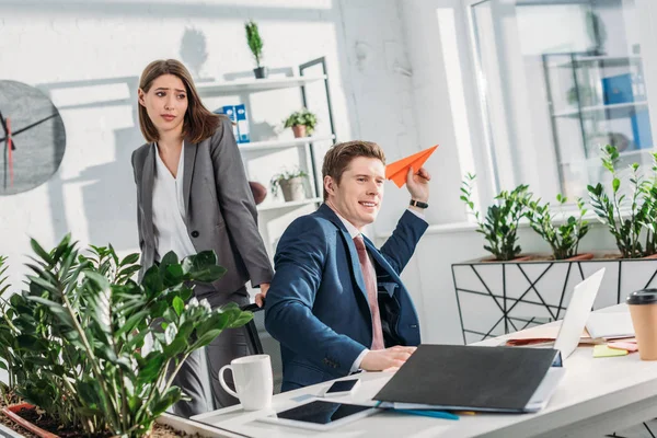 Attractive businesswoman standing and looking at cheerful coworker holding paper plane in office — Stock Photo