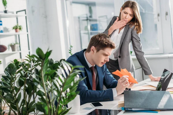 Tired businesswoman standing and yawning near coworker holding paper plane in office — Stock Photo