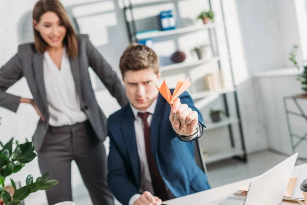 Selective focus of paper plane in hand of businessman sitting in office with female coworker on background — Stock Photo