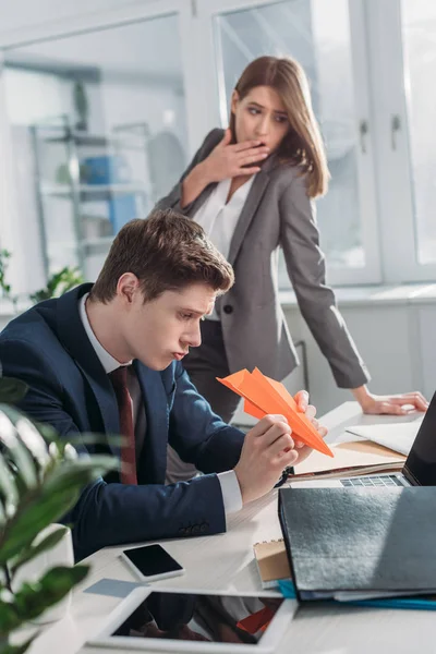 Exhausted businesswoman standing and yawning near coworker holding paper plane in office — Stock Photo