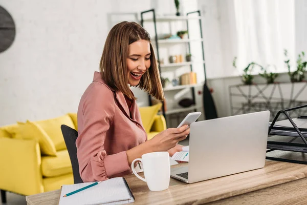 Mujer alegre sosteniendo teléfono inteligente cerca de la computadora portátil en casa - foto de stock