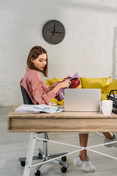 Mulher atraente segurando revista perto de laptop em casa — Fotografia de Stock