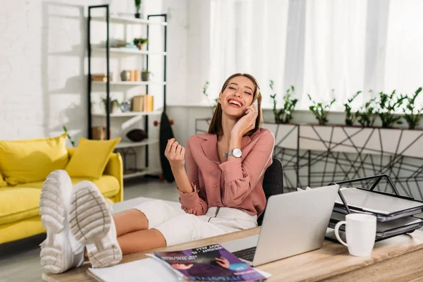 Selektiver Fokus der glücklichen Frau, die auf dem Smartphone spricht, während sie neben dem Laptop sitzt — Stockfoto