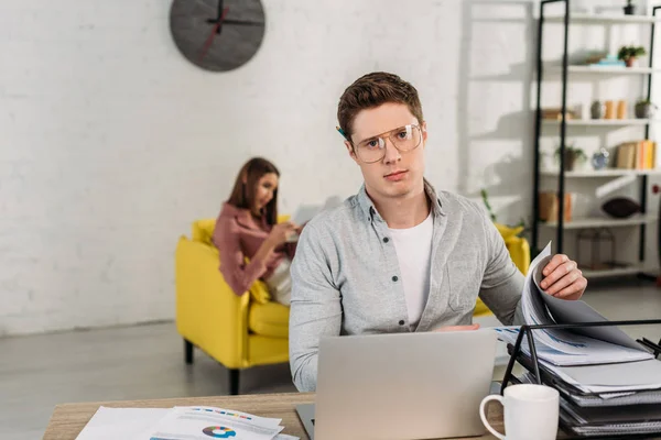 Handsome man in glasses sitting at desk near laptop with girlfriend on background — Stock Photo