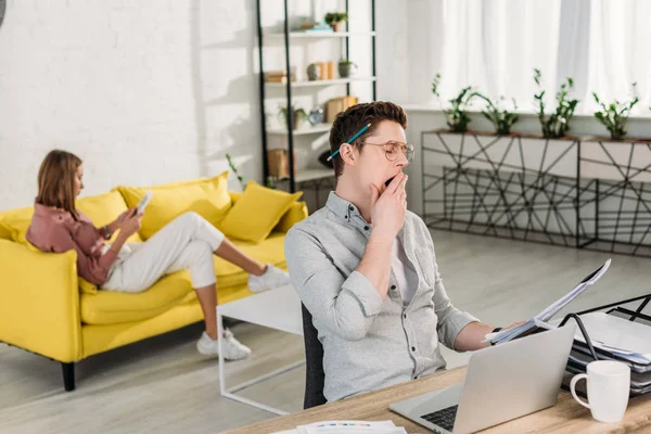 Müder Mann gähnt, während er Dokumente in der Nähe von Laptop mit Freundin im Hintergrund hält — Stockfoto