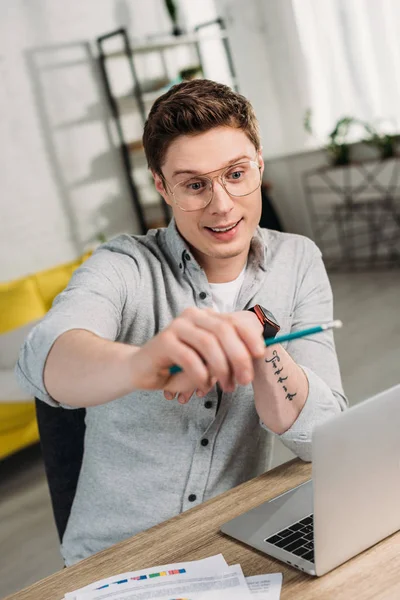Selective focus of cheerful man in glasses holding pencil and sitting near laptop at home — Stock Photo