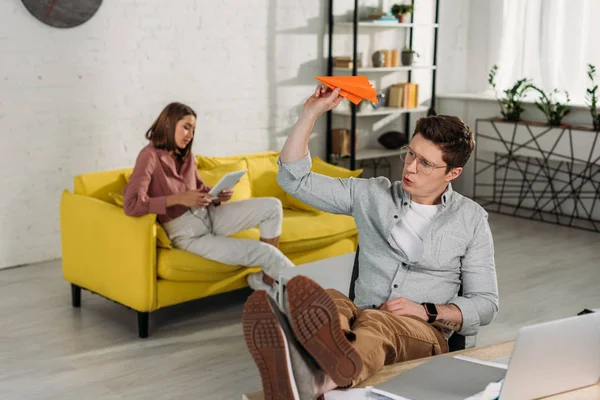 Selective focus of man playing with paper plane near laptop with girlfriend on background — Stock Photo