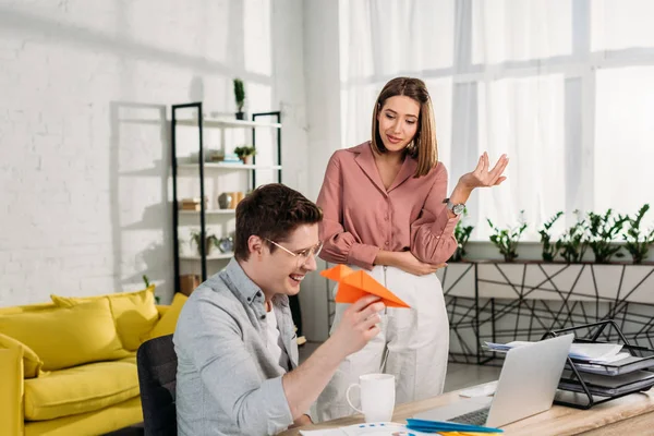 Woman gesturing near cheerful boyfriend playing with paper plane near laptop at home — Stock Photo