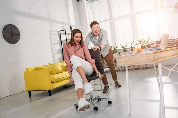 Cheerful man smiling while pushing chair of happy girlfriend — Stock Photo