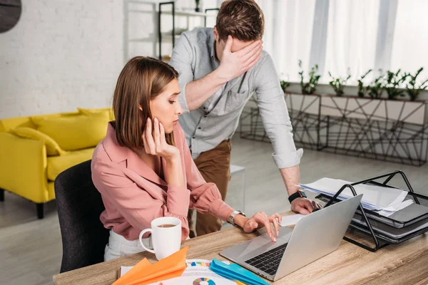 Man standing and covering eyes near tired girlfriend sitting and looking at laptop — Stock Photo