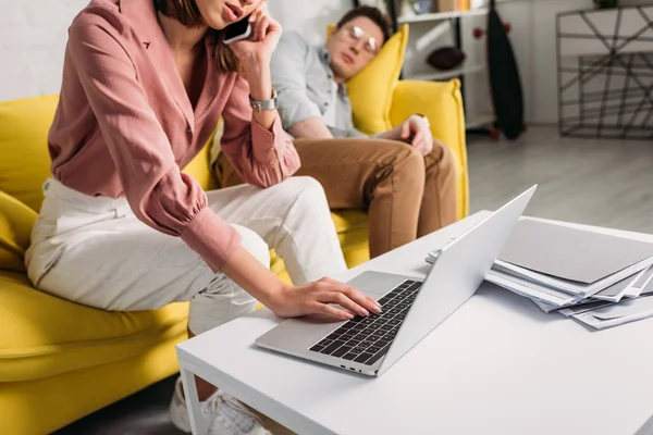 Vista recortada de la mujer hablando en el teléfono inteligente y escribiendo en el ordenador portátil cerca de novio en el sofá - foto de stock