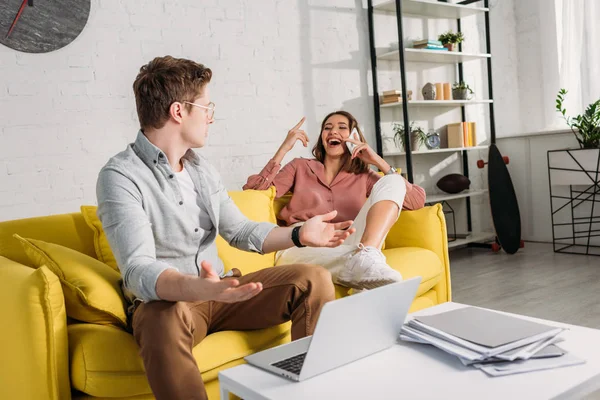 Selective focus of man in glasses gesturing near laptop while looking at girlfriend talking on smartphone — Stock Photo