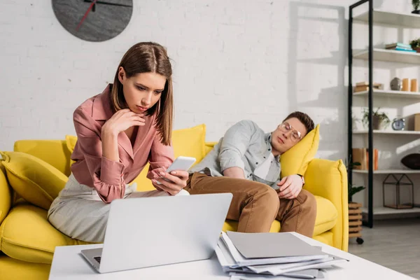 Atractiva mujer sosteniendo teléfono inteligente cerca novio durmiendo en sofá - foto de stock