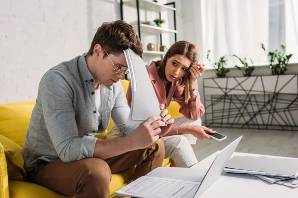 Atractiva mujer mirando guapo cansado novio en gafas celebración carpeta - foto de stock