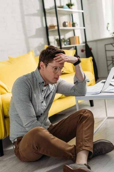 Tired man in glasses sitting on floor and holding head at home — Stock Photo