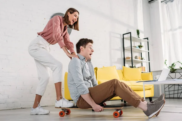 Petite amie joyeuse poussant skateboard de petit ami dans des lunettes à la maison — Photo de stock