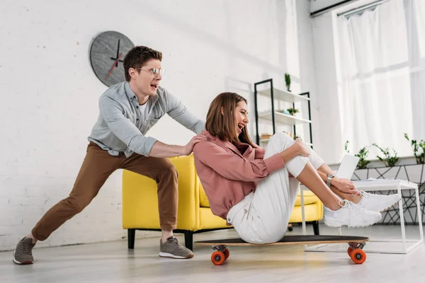 Petit ami joyeux poussant skateboard de petite amie à la maison — Photo de stock