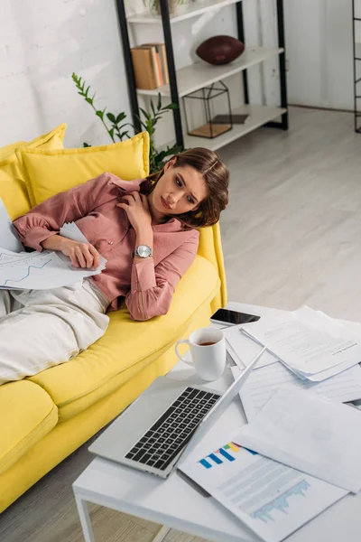 Mujer cansada acostada en el sofá con documentos y mirando la taza con la bebida cerca de la computadora portátil - foto de stock