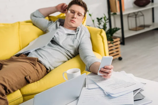 Selektive Fokussierung des Laptops mit Mann, der Smartphone nutzt und auf Sofa im Hintergrund liegt — Stockfoto