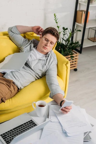 Uomo in occhiali utilizzando smartphone e sdraiato sul divano vicino ai documenti — Foto stock