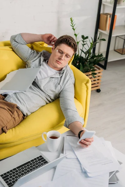 Mann mit Brille benutzt Smartphone und liegt auf Sofa in der Nähe von Dokumenten — Stockfoto