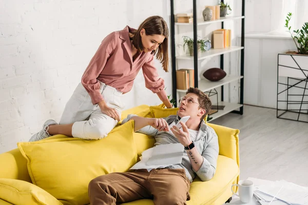 Attractive woman looking at boyfriend with smartphone lying on sofa — Stock Photo