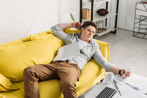 Uomo utilizzando smartphone mentre sdraiato sul divano vicino ai documenti — Foto stock