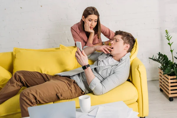 Attractive woman looking at  tired boyfriend using smartphone and lying on sofa — Stock Photo