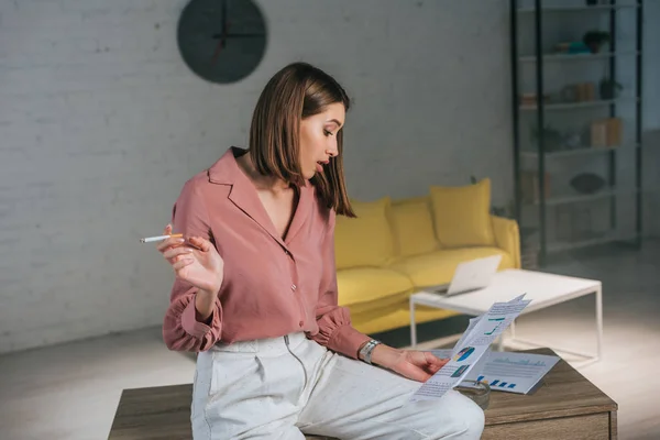 Mujer atractiva sosteniendo el cigarrillo y mirando gráficos y gráficos en casa - foto de stock