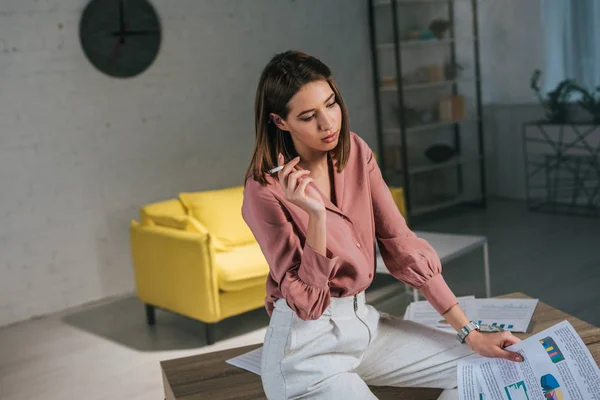 Pensive woman holding cigarette and holding charts and graphs at home — Stock Photo