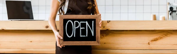 Cropped view of cashier near wooden bar counter holding chalkboard with open lettering in coffee house — Stock Photo