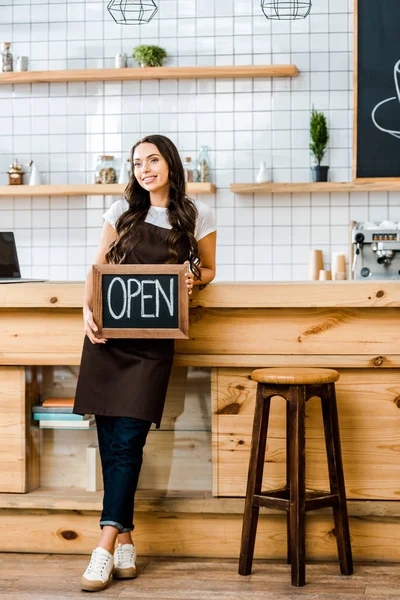 Beautiful cashier standing near wooden bar counter and holding chalkboard with open lettering in coffee house — Stock Photo