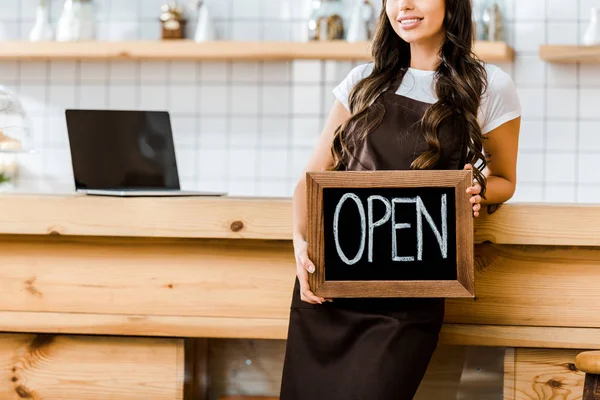 Ausgeschnittene Ansicht der Kassiererin, die in der Nähe der hölzernen Theke steht und eine Tafel mit offenem Schriftzug im Kaffeehaus hält — Stockfoto