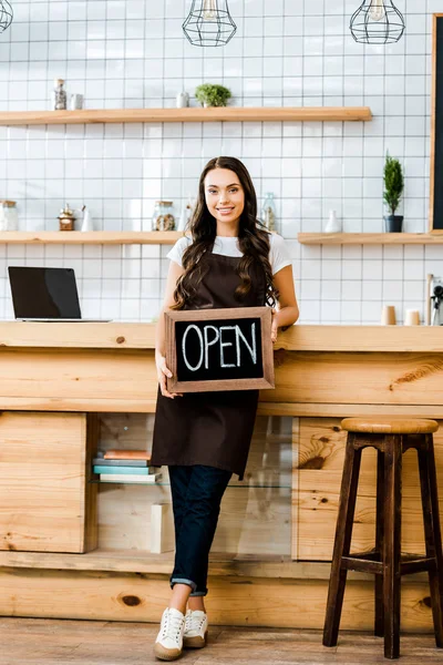 Beautiful cashier in apron standing near wooden bar counter and holding chalkboard with open lettering in coffee house — Stock Photo
