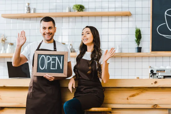 Caissier et barista salutation et tenue tableau noir avec lettrage ouvert près comptoir de bar en bois dans le café — Photo de stock