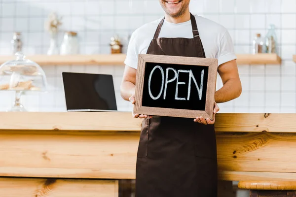 Vista recortada de cajero en delantal de pie cerca del mostrador de bar de madera y la celebración de pizarra con letras abiertas en la cafetería - foto de stock