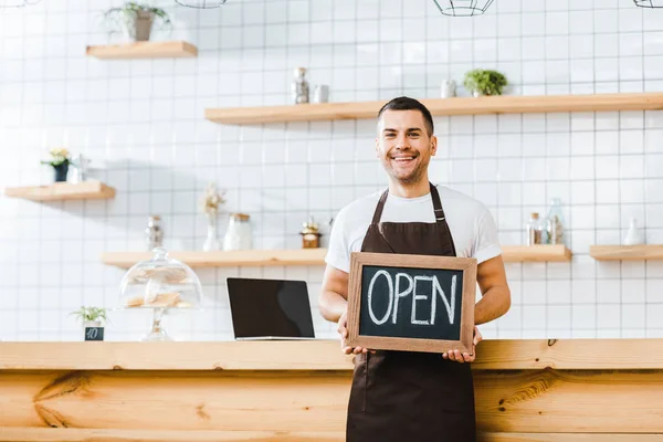 Bel cassiere in grembiule marrone in piedi vicino al bancone del bar in legno e contenente lavagna con scritte aperte in caffetteria — Foto stock