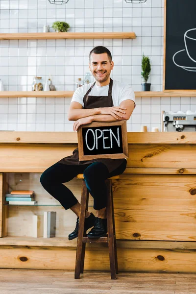 Cajero en delantal sentado en la silla cerca del mostrador de bar de madera y la celebración de pizarra con letras abiertas en la cafetería - foto de stock