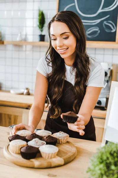Selective focus of attractive waitress standing behind bar counter and putting cupcakes on wooden tray in coffee house — Stock Photo