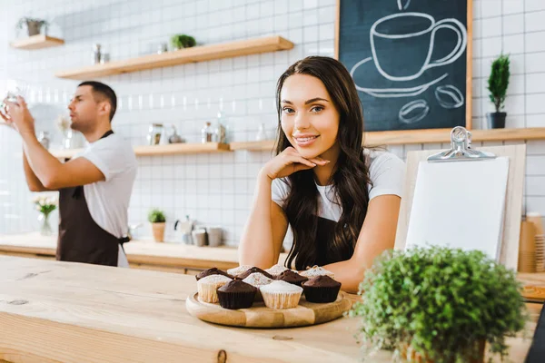 Приваблива брюнетка офіціантка стоїть за барною стійкою з кексами на дерев'яному лотку бариста тримає чашку в кав'ярні — стокове фото