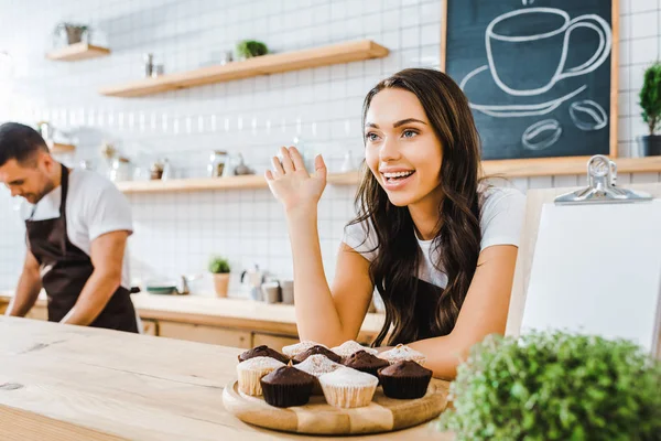 Selective focus of attractive brunette waitress standing behind bar counter with cupcakes and waving wile cashier working in coffee house — Stock Photo