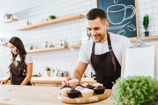 Selective focus of waiter standing behind bar counter with cupcakes wile cashier working in coffee house — Stock Photo