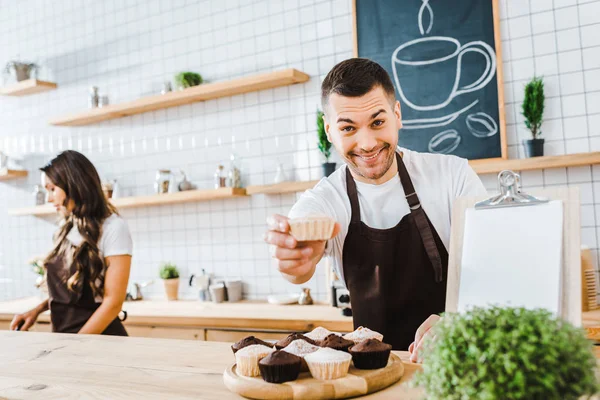 Selective focus of handsome barista giving cupcake wile attractive brunette cashier working in coffee house — Stock Photo